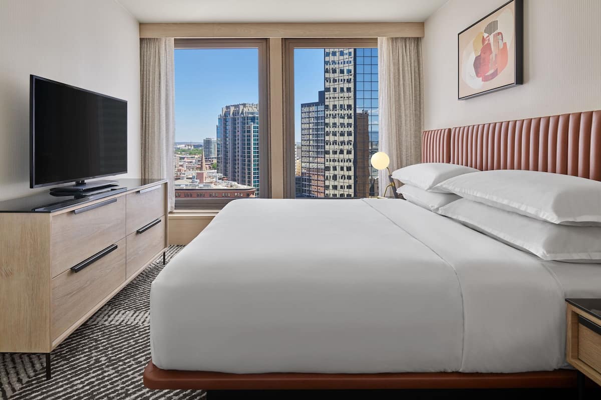A hotel room with a large bed, a flat-screen TV, a wooden dresser, and a city view through large windows.
