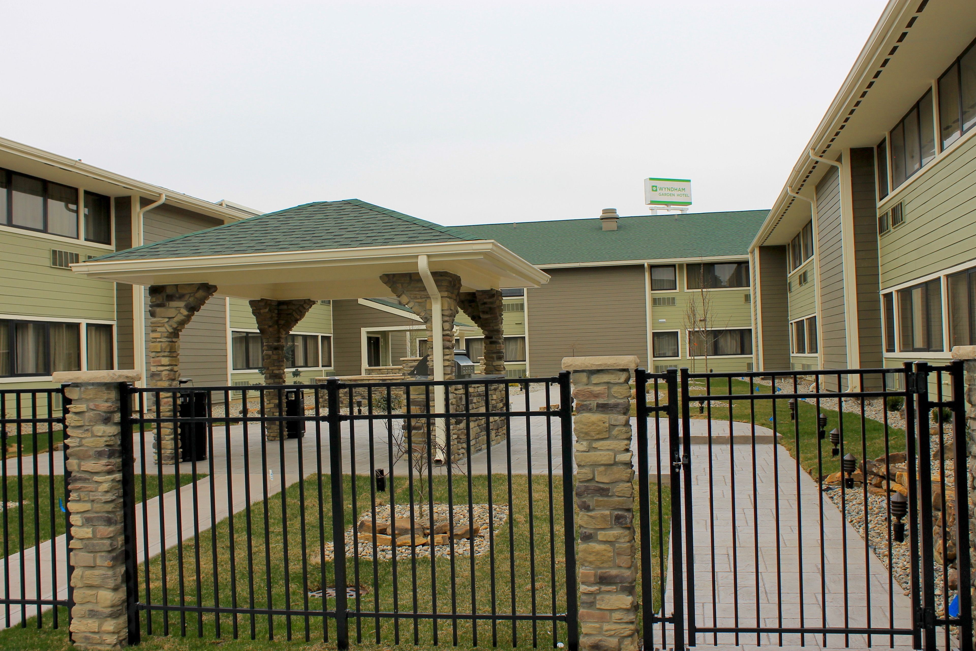 A gated courtyard with a covered seating area, stone pillars, and a pathway leading to a building with a green roof.
