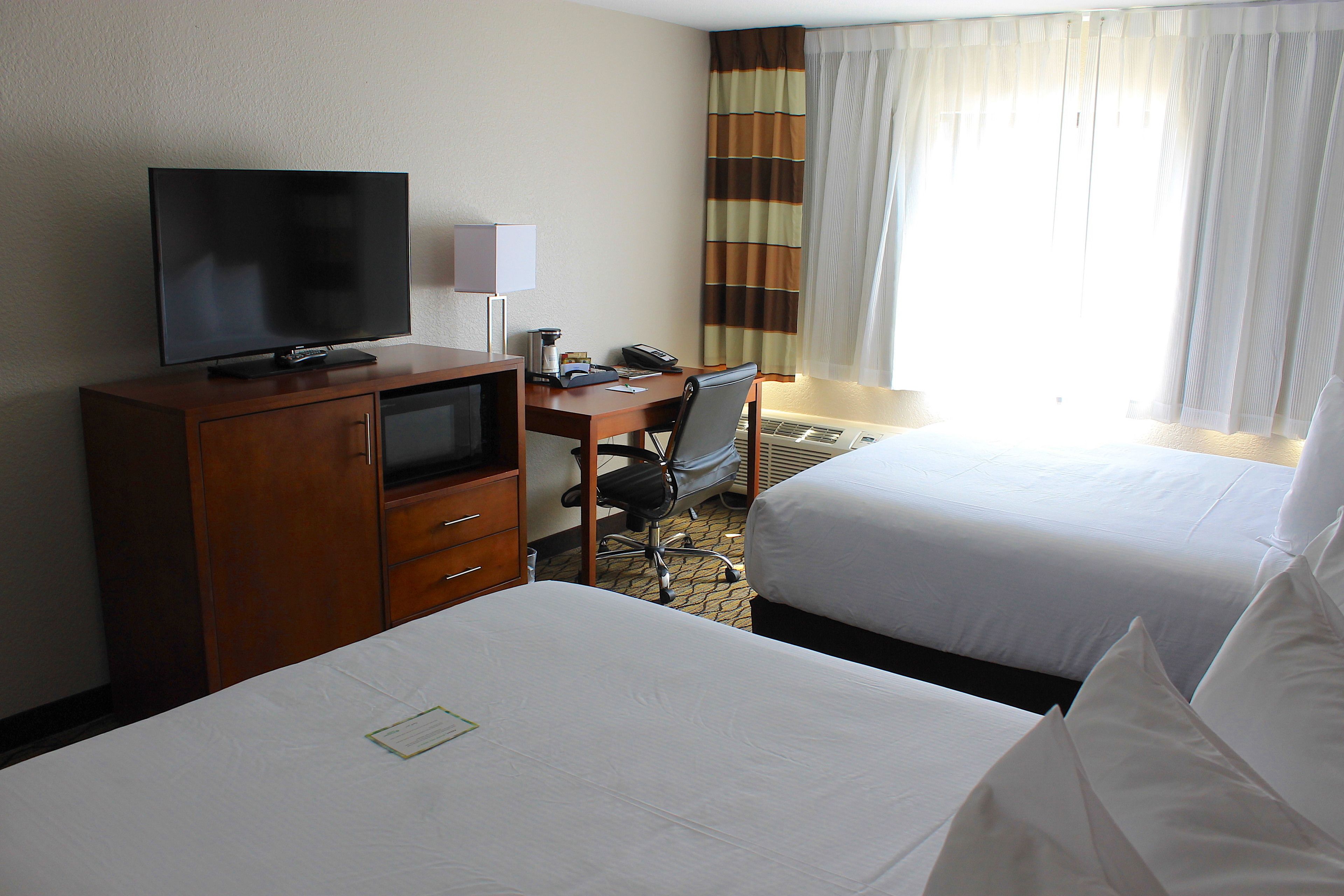 Habitación de hotel con televisión sobre un mueble de madera, un escritorio con lámpara, una silla y dos camas.