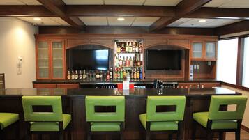 Una zona de bar con un mostrador de madera, taburetes verdes y una estantería llena de botellas de licor.