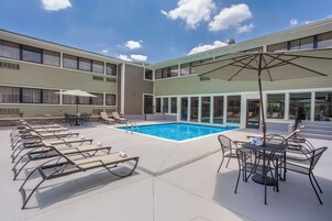 Un área de piscina con sillones de descanso y sombrillas, rodeada por un edificio con grandes ventanales y una piscina.