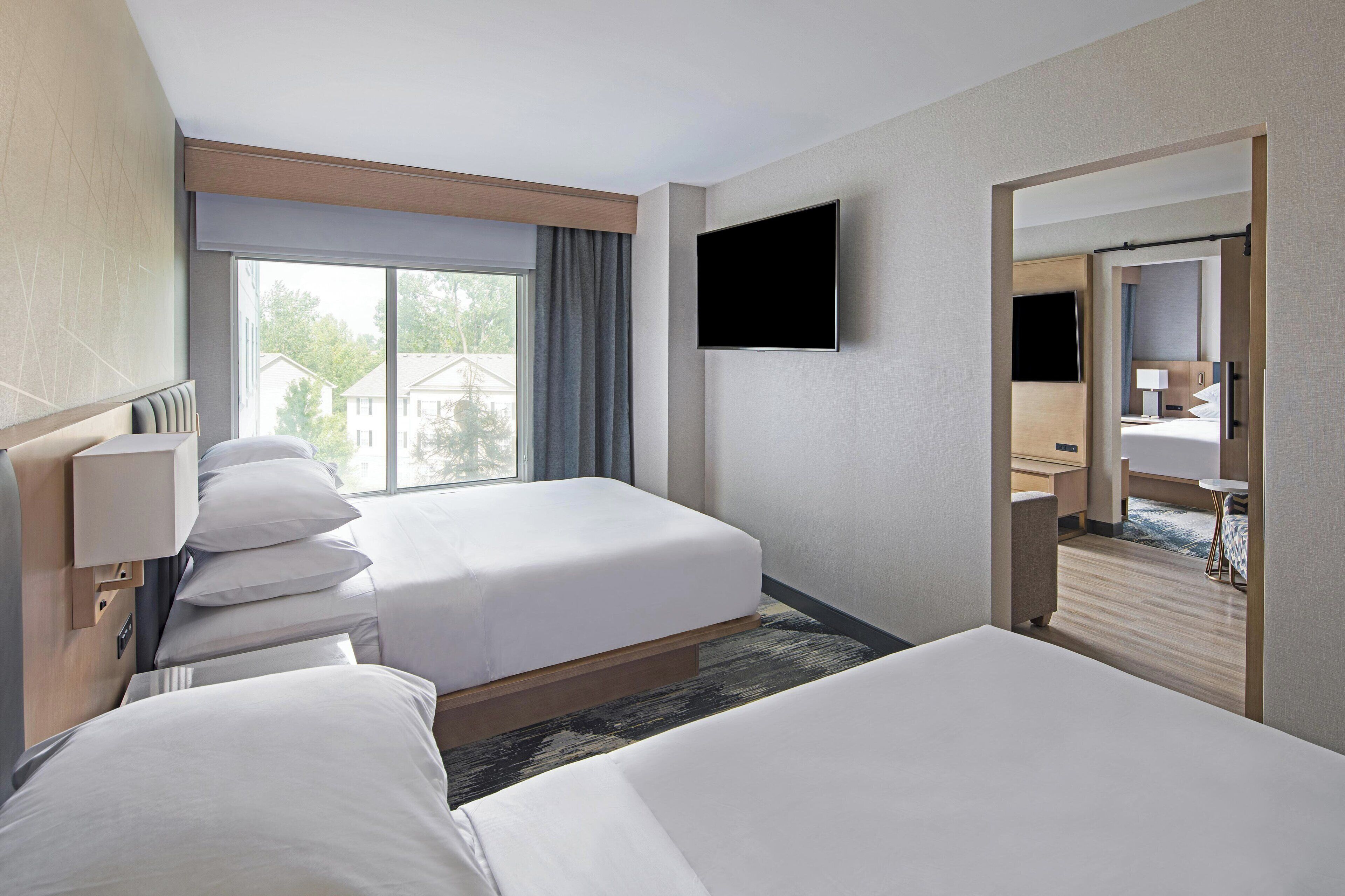 A hotel room with two beds, a TV mounted on the wall, a window with curtains, and a view of buildings outside.