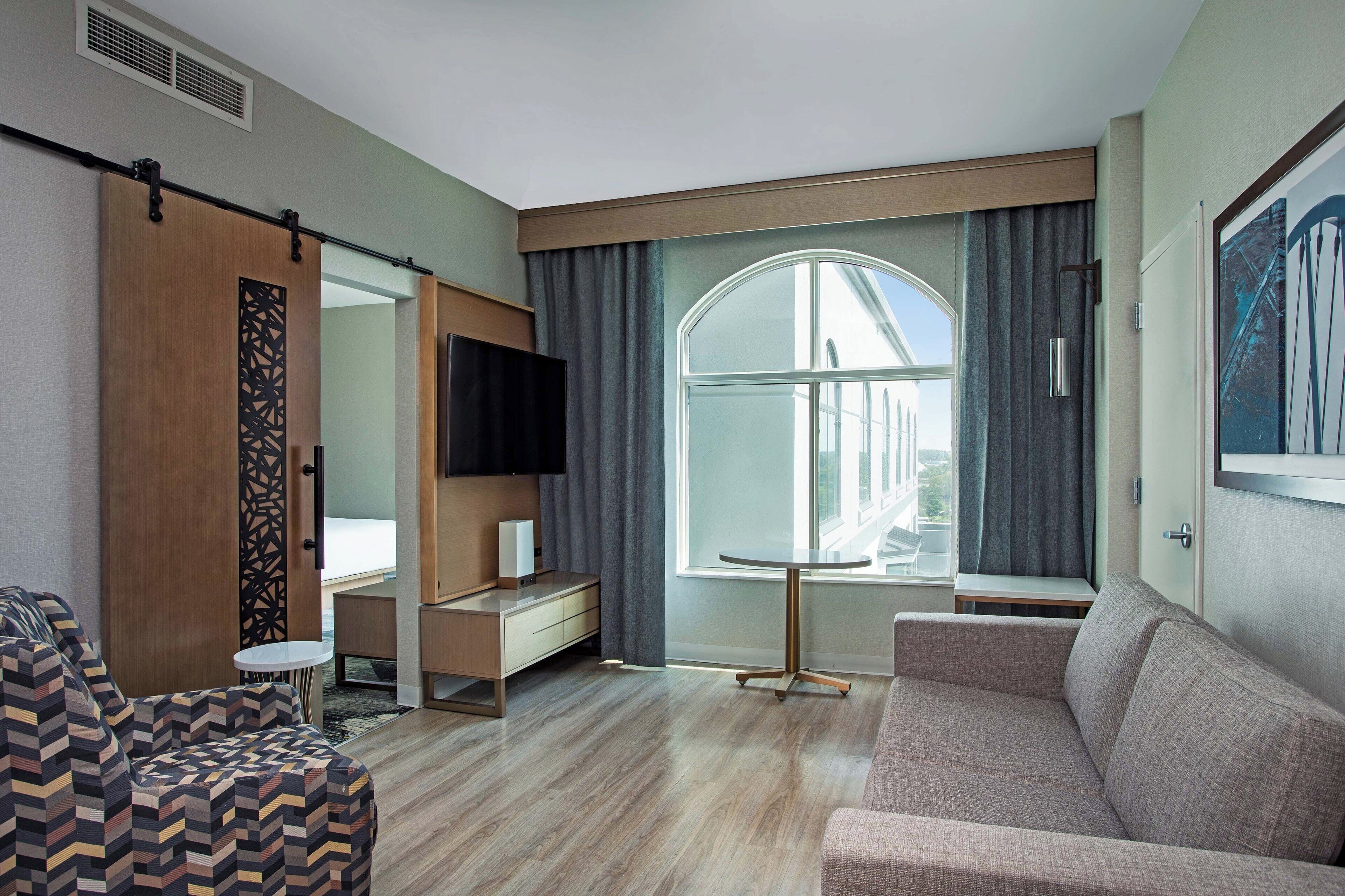 A modern hotel room with a large window, a sofa, a TV, and a small table.