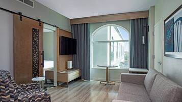 A modern hotel room with a large window, a sofa, a TV, and a small table.
