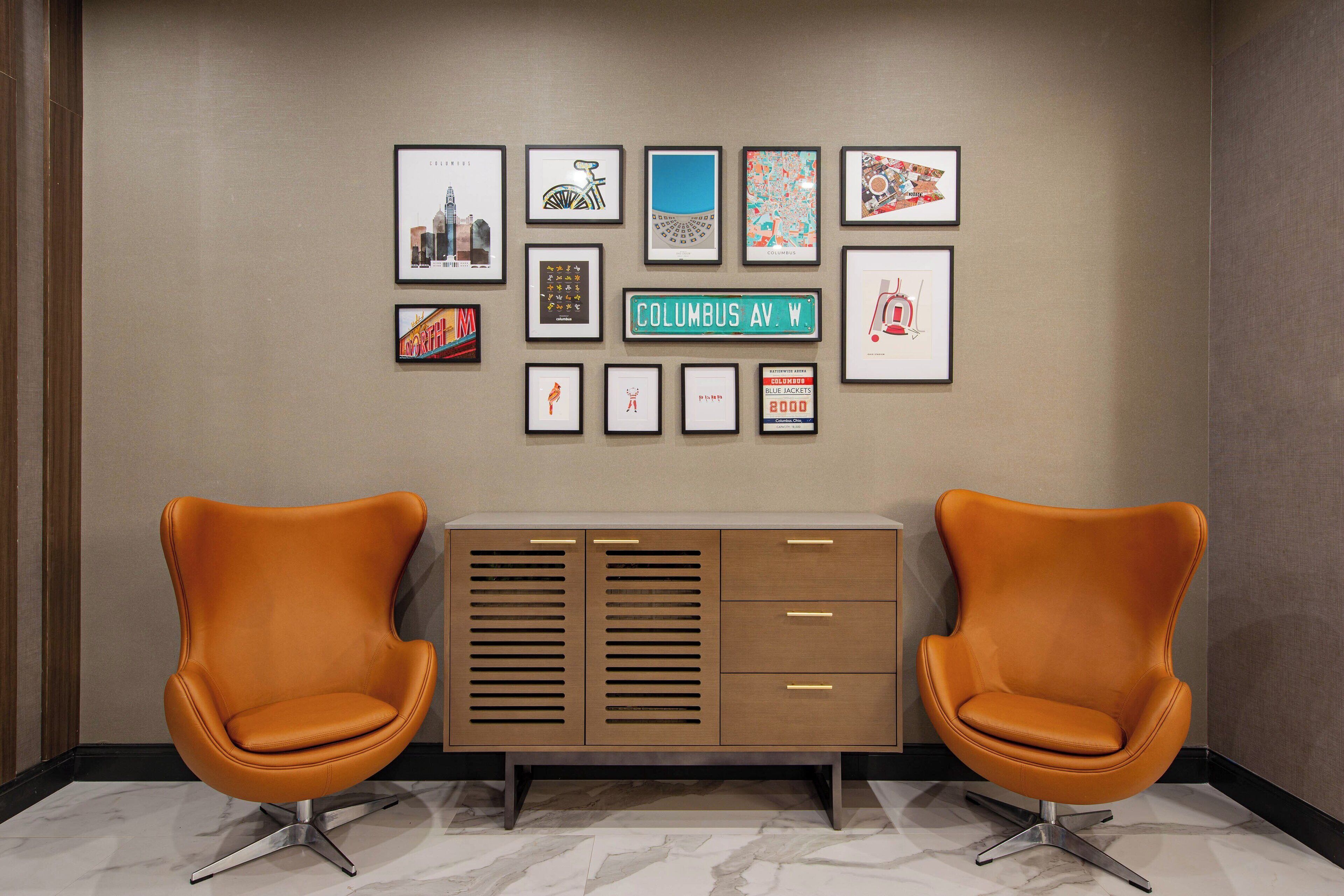 A lobby with two orange chairs, a wooden cabinet with drawers, and a wall decorated with framed pictures.