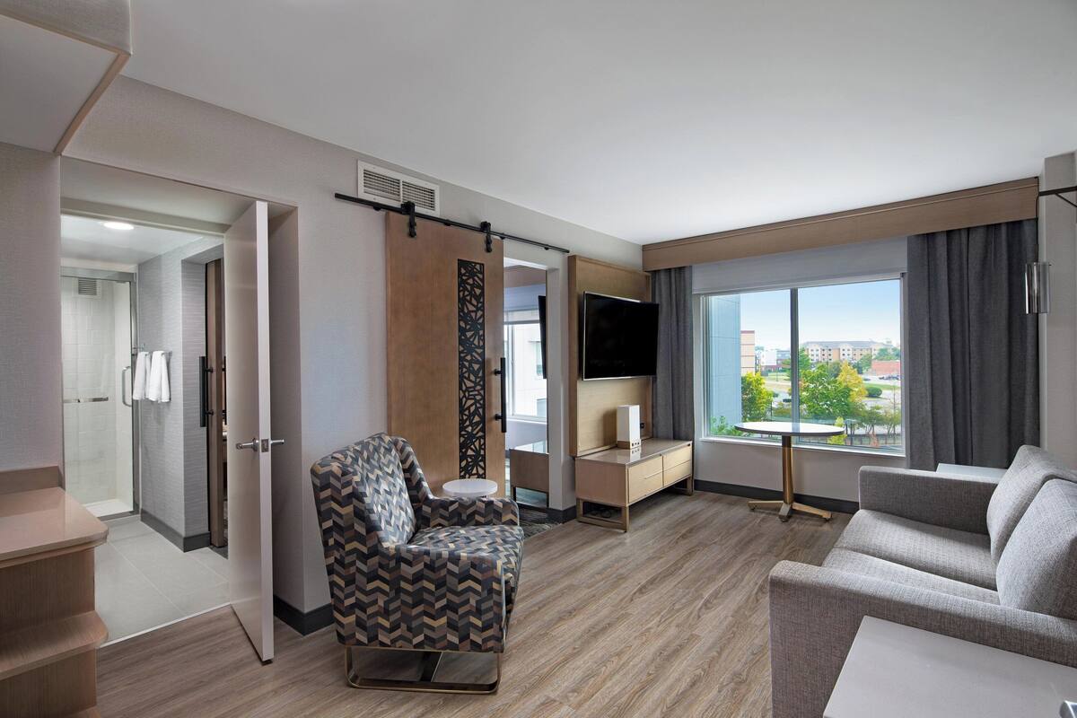 A modern hotel room with a sofa, armchair, TV, and a view of the city.