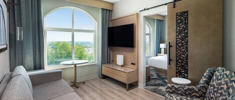 Une chambre d’hôtel avec une grande fenêtre, un canapé, un fauteuil, un bureau, une télévision et un lit.