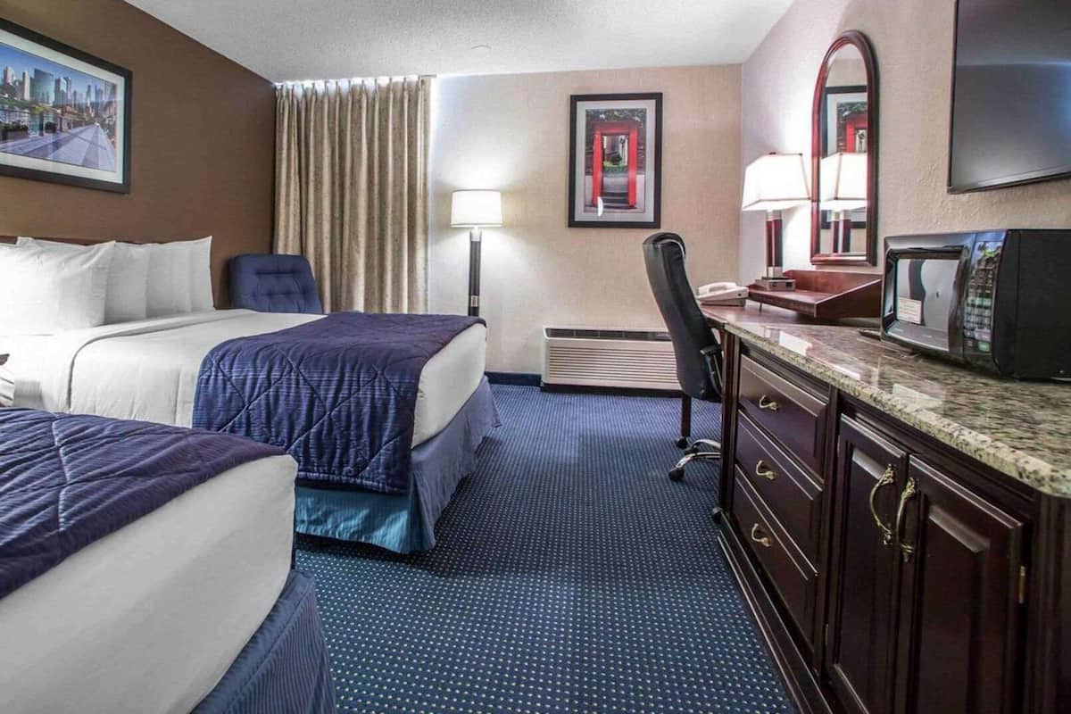 A hotel room with two beds, a desk with a chair, a desk lamp, a telephone, a microwave, and a television.