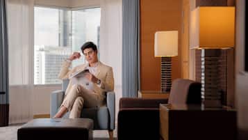 A man sitting in a modern hotel room with a large window, a lamp, and a stack of books.