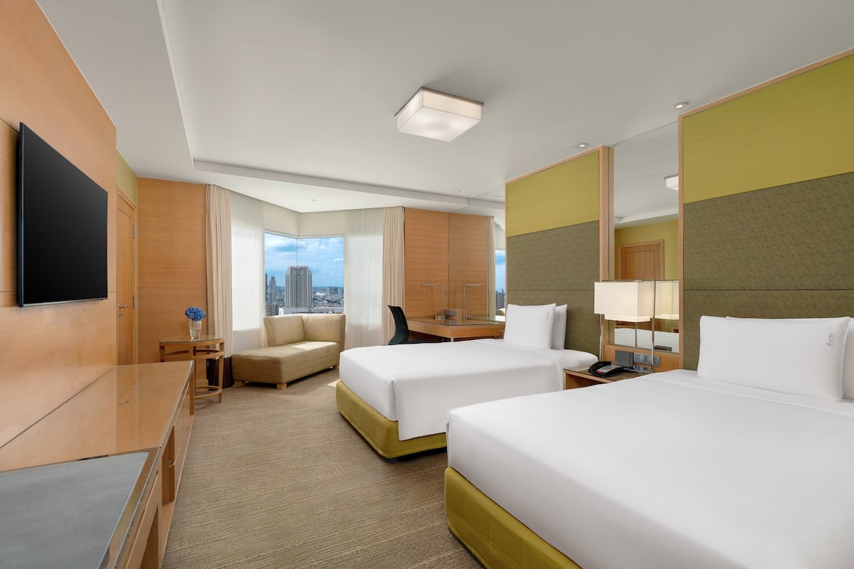 Habitación de hotel con dos camas, un escritorio, una silla, un televisor y vistas a la ciudad.