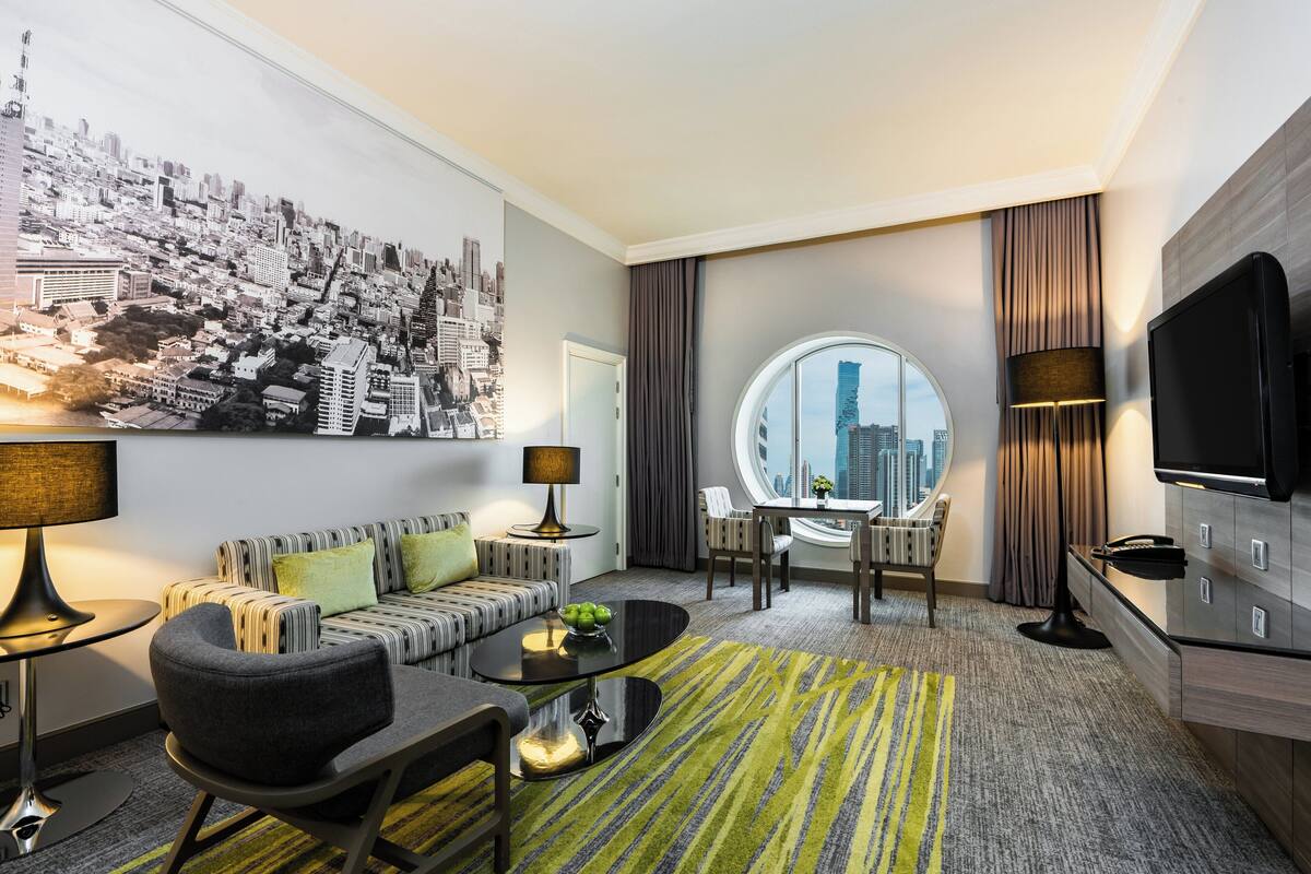 Una habitación de hotel moderna con un mural de la ciudad, un ventanal redondo que ofrece vistas urbanas, un sofá, una mesa de centro y un televisor.