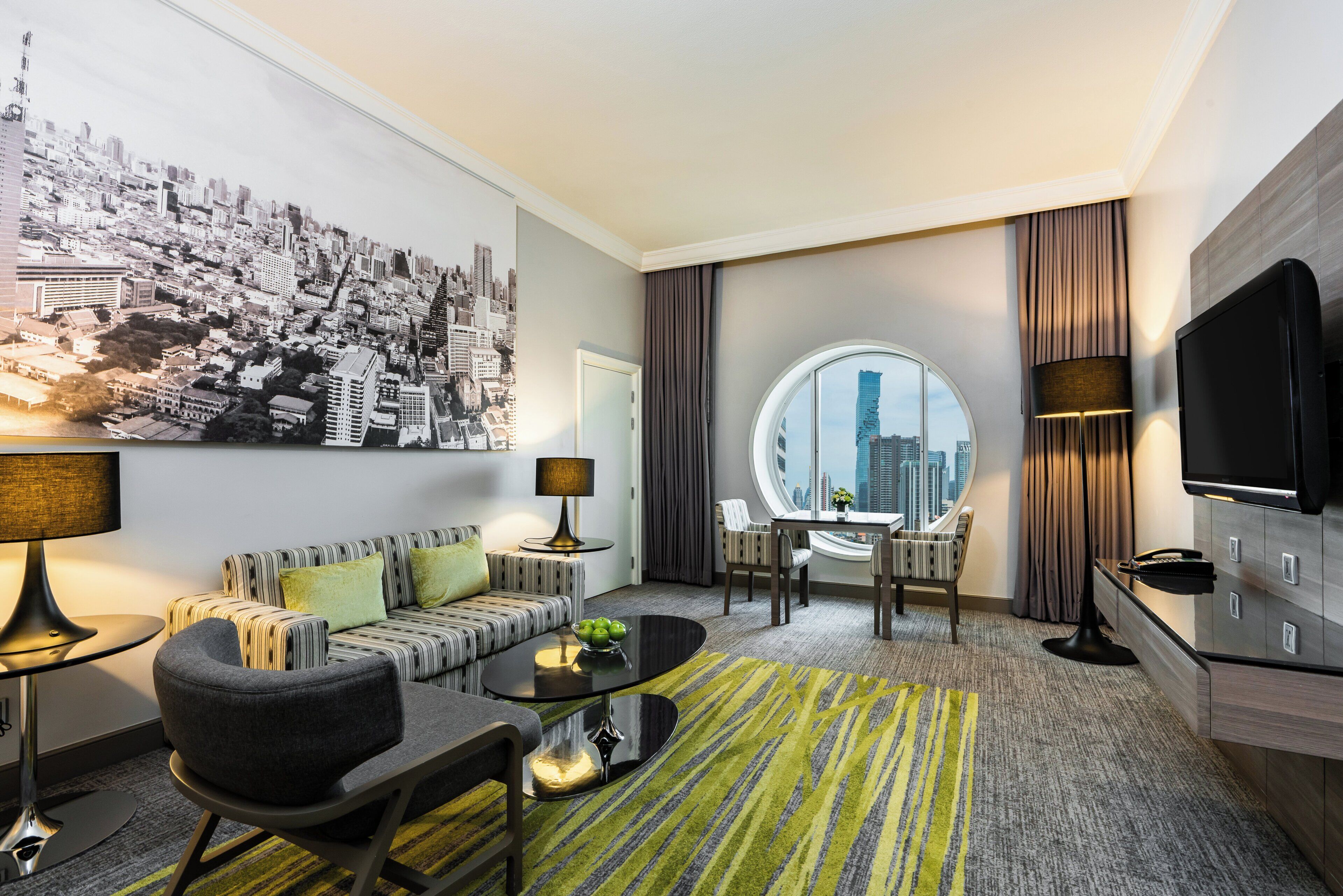 A modern hotel room with a cityscape mural, a round window with a city view, a sofa, a coffee table, and a TV.