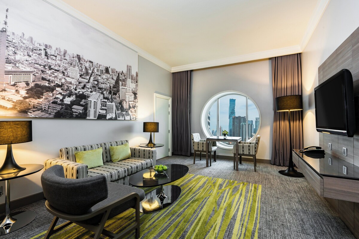 A modern hotel room with a cityscape mural, a round window with a city view, a sofa, a coffee table, and a TV.