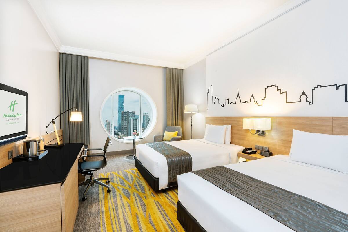 Habitación de hotel con dos camas, un escritorio, una silla, un espejo redondo y vistas a la silueta urbana.