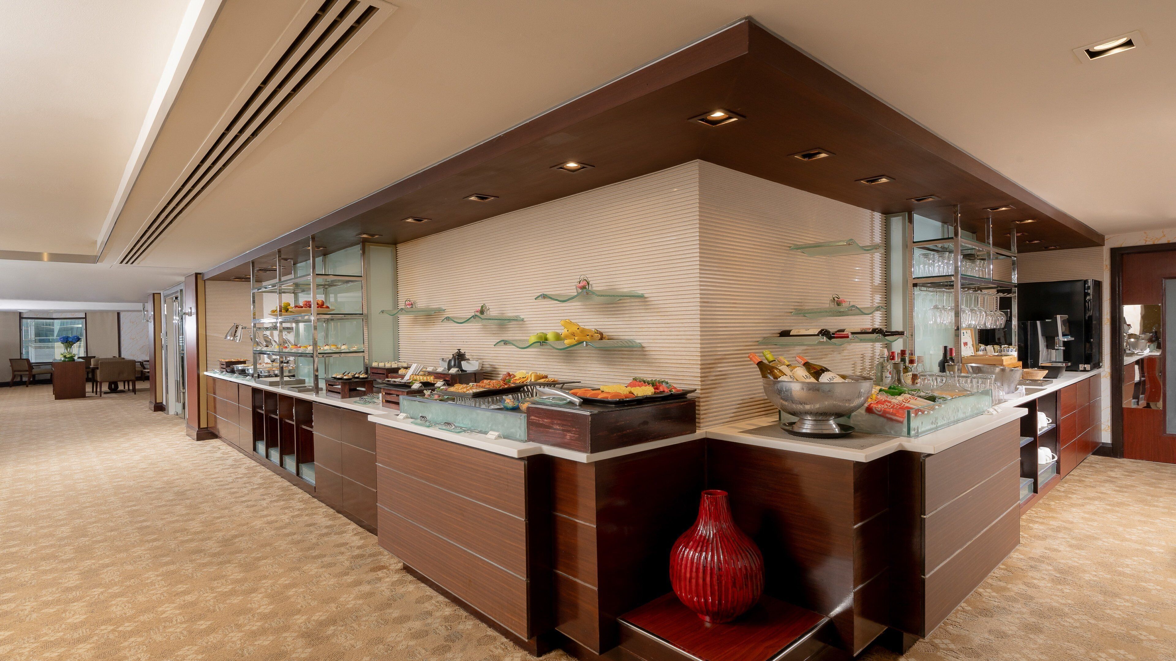Eine moderne Hotelhalle mit einem Buffetbereich, der Glasvitrinen, eine rote Vase und eine Kaffeemaschine zeigt.