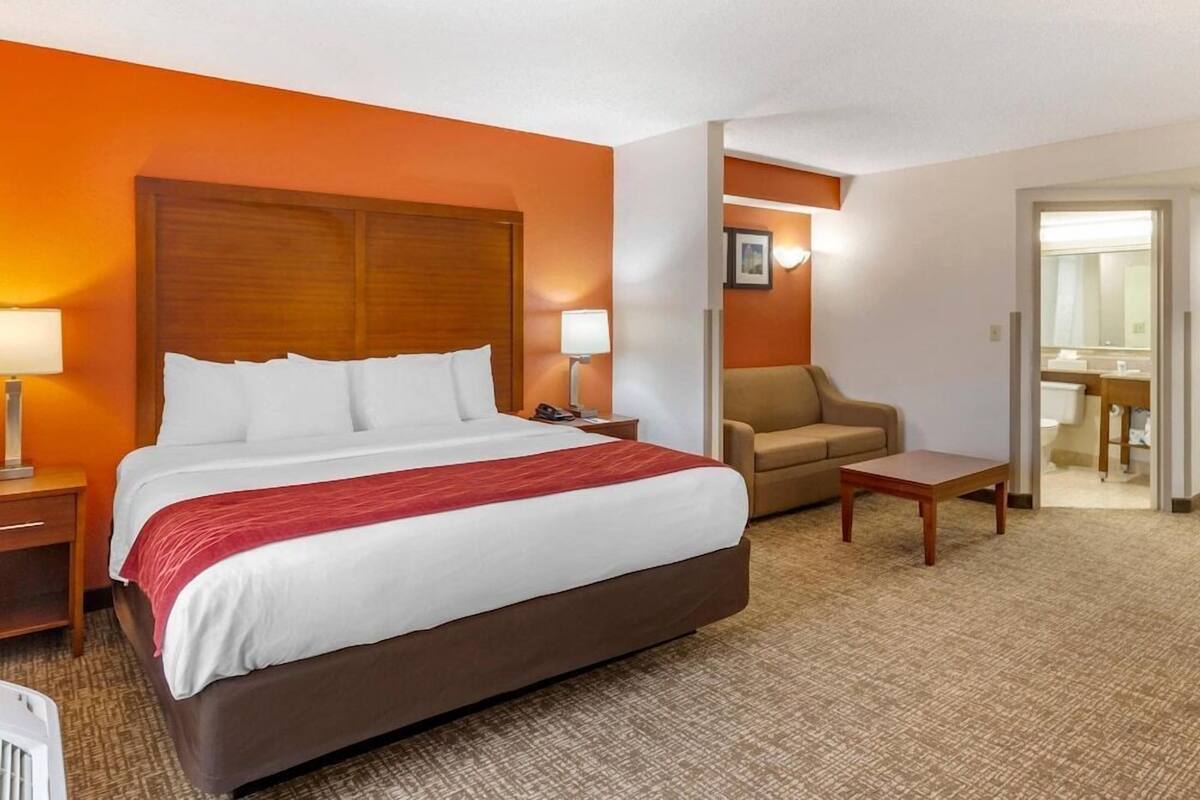 Une chambre d’hôtel avec un grand lit, une chaise, une petite table et une salle de bain visible à travers une porte ouverte.