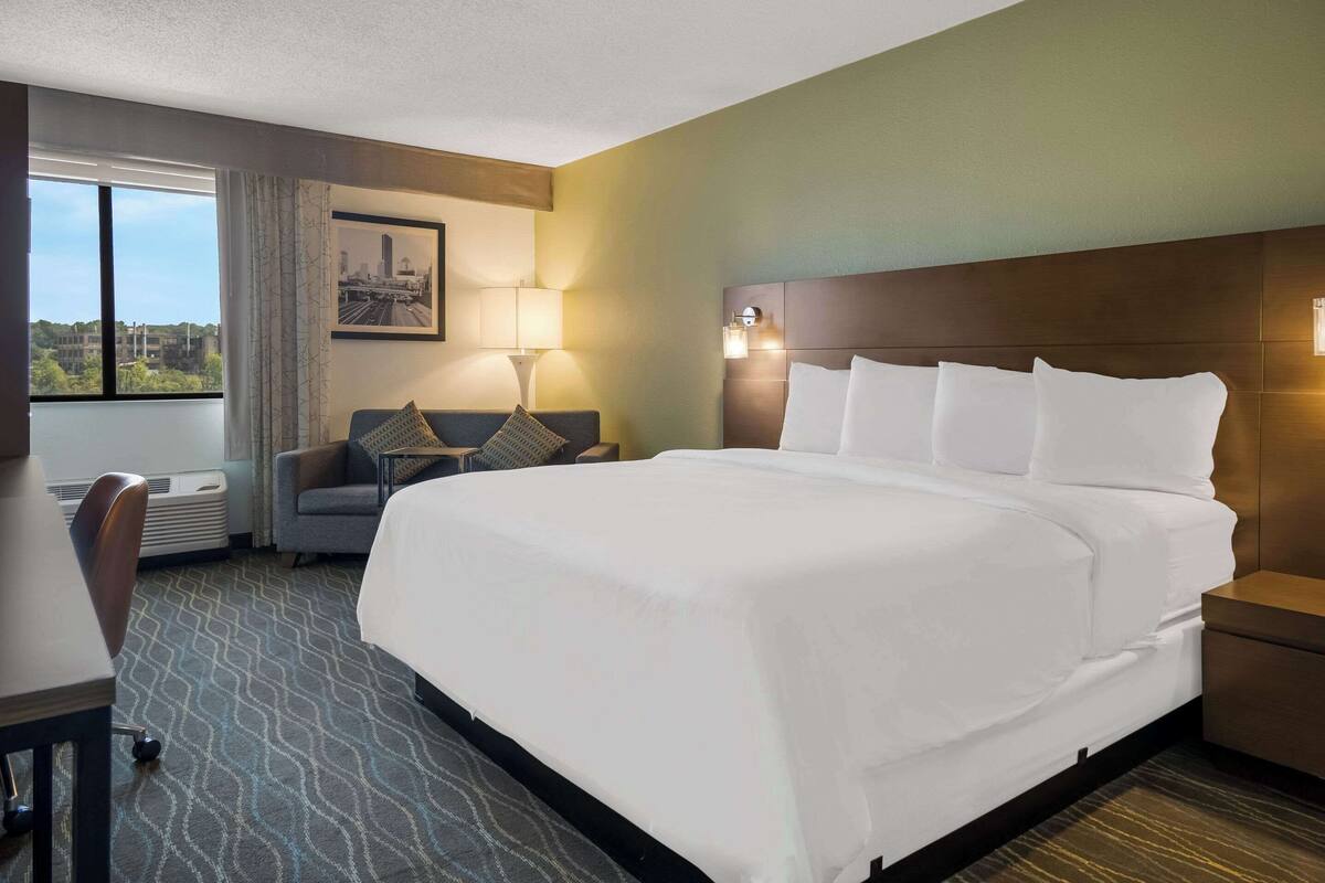 A hotel room with a large bed, a desk, a chair, a window with a city view, and a wall-mounted lamp.