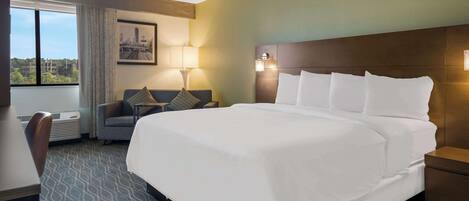 A hotel room with a large bed, a desk, a chair, a window with a city view, and a wall-mounted lamp.