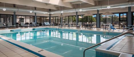 A large indoor swimming pool with clear water, surrounded by lounge chairs and tables, and large windows offering views of the outdoors.
