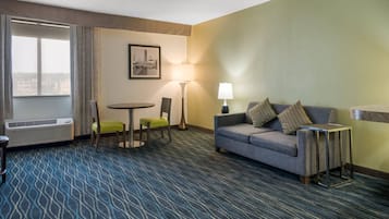 Habitación de hotel con un sofá gris, una mesita, una silla, una lámpara y un cuadro en la pared.
