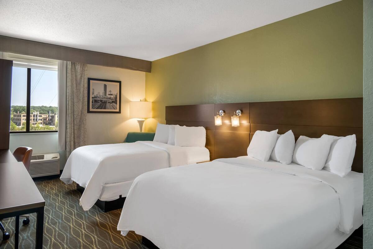 A hotel room with two beds, a desk, and a window with a city view.