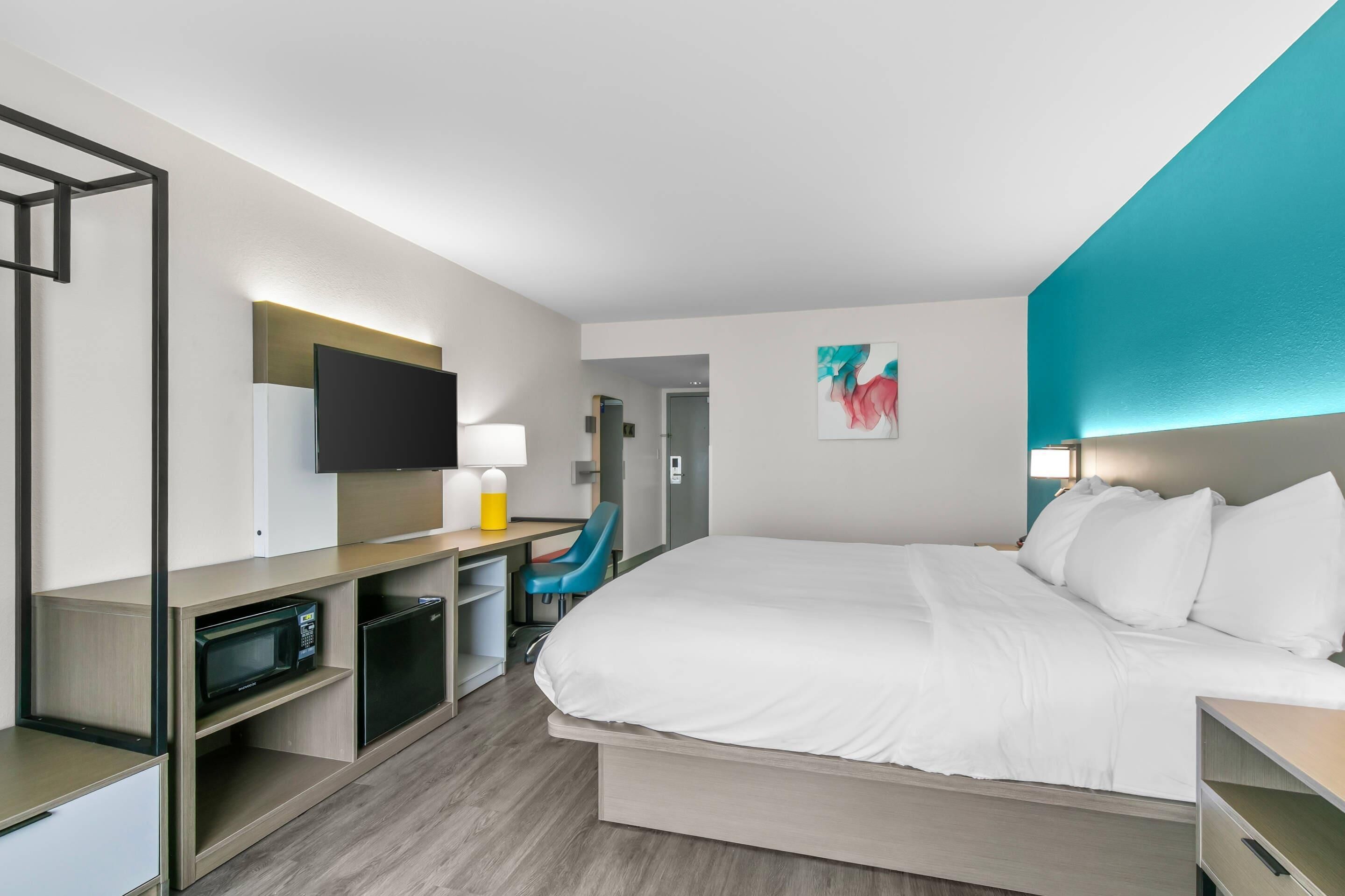 A hotel room with a bed, desk, chair, TV, and artwork on the wall.