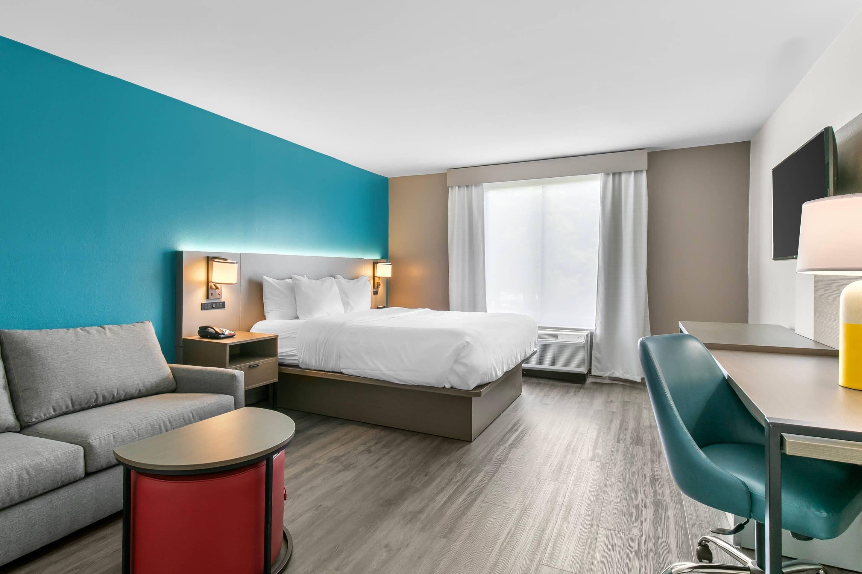 A hotel room with a bed, a desk, a chair, a sofa, a bedside table, and a window with curtains.