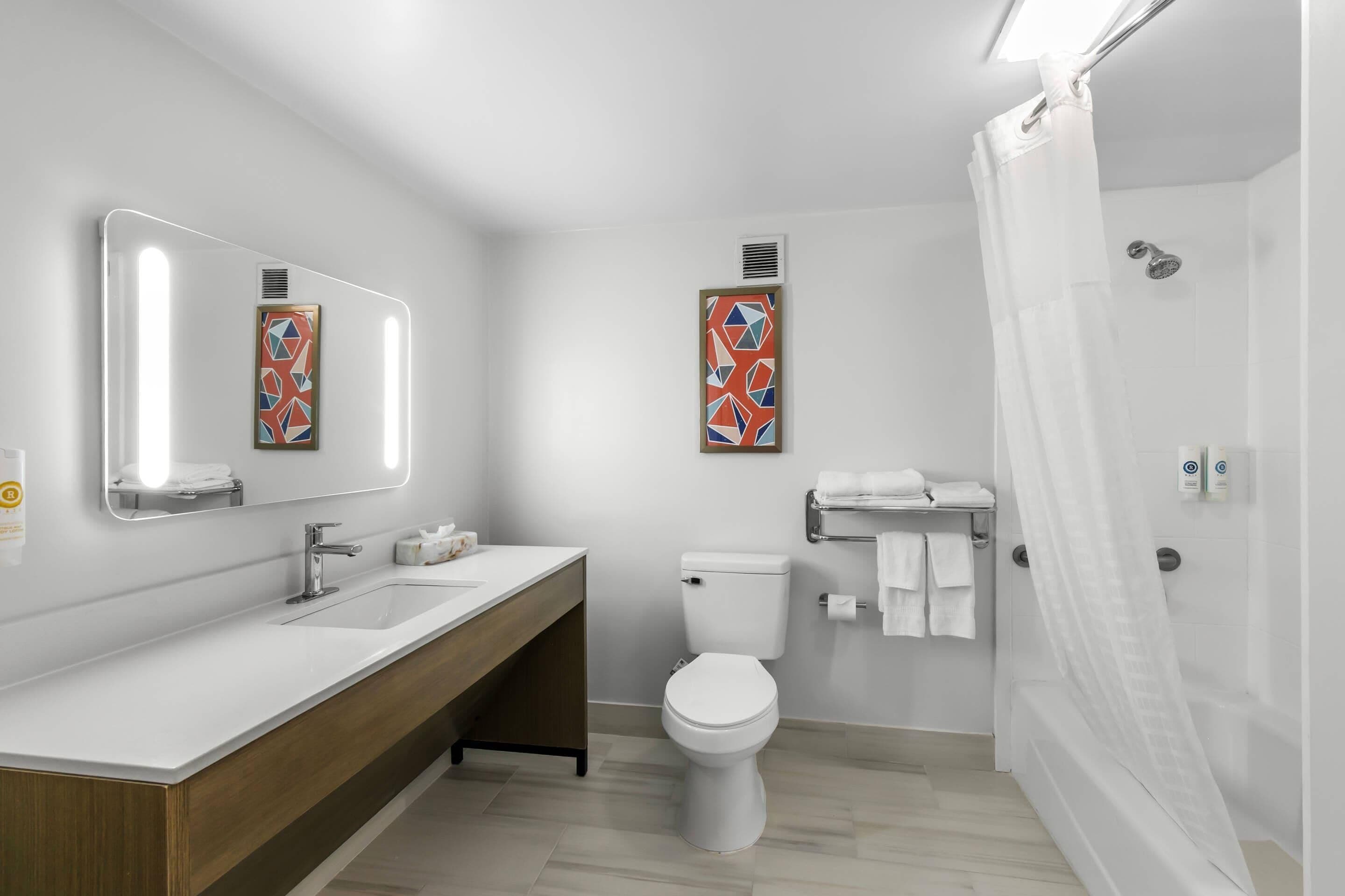 Uma casa de banho moderna com lavatório, sanita e chuveiro, todos em branco. Possui duas espelhas fixadas na parede e um porta-toalhas.