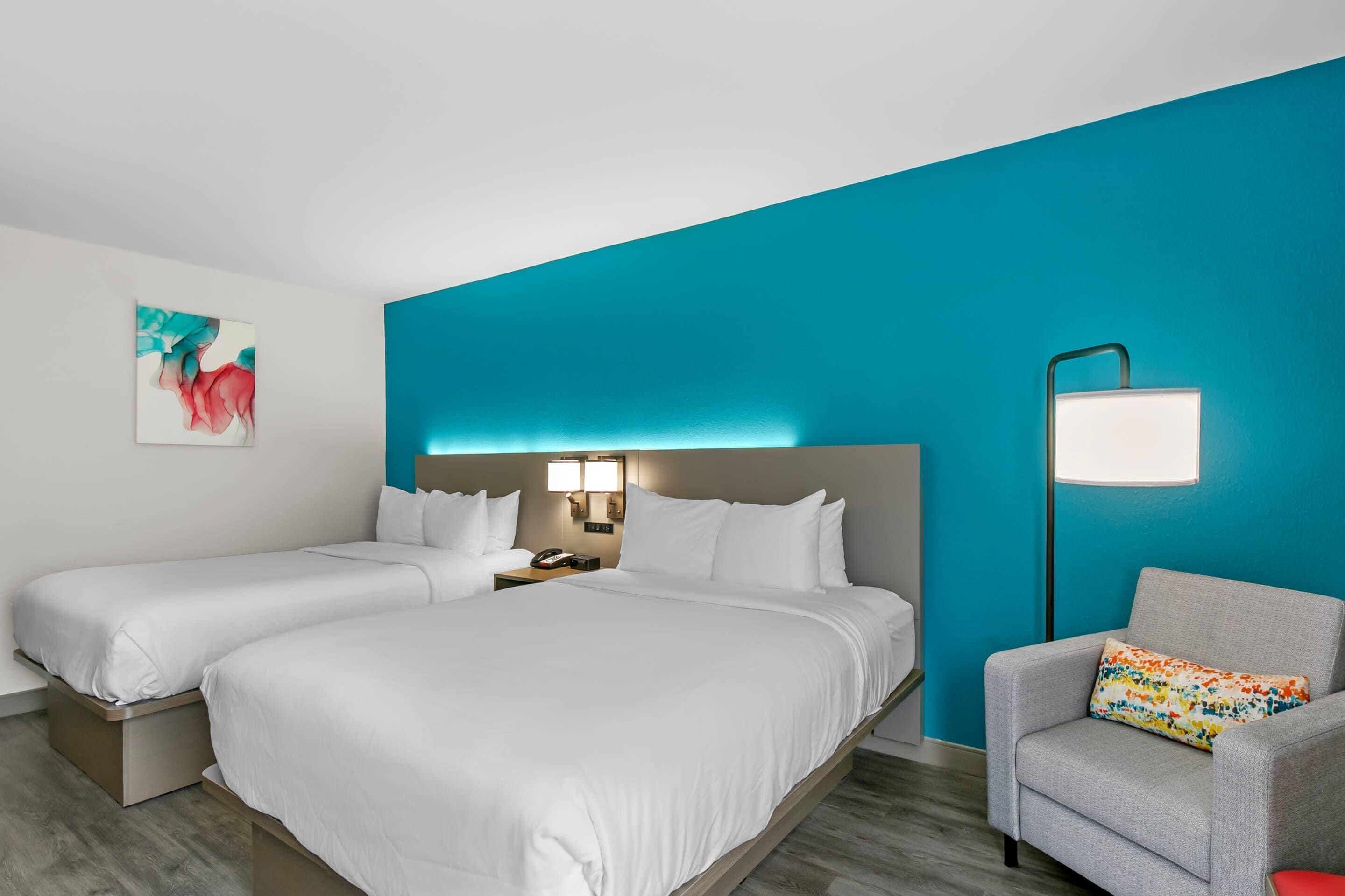 A hotel room with two beds, a nightstand with a phone, a lamp, and a chair with a colorful pillow.