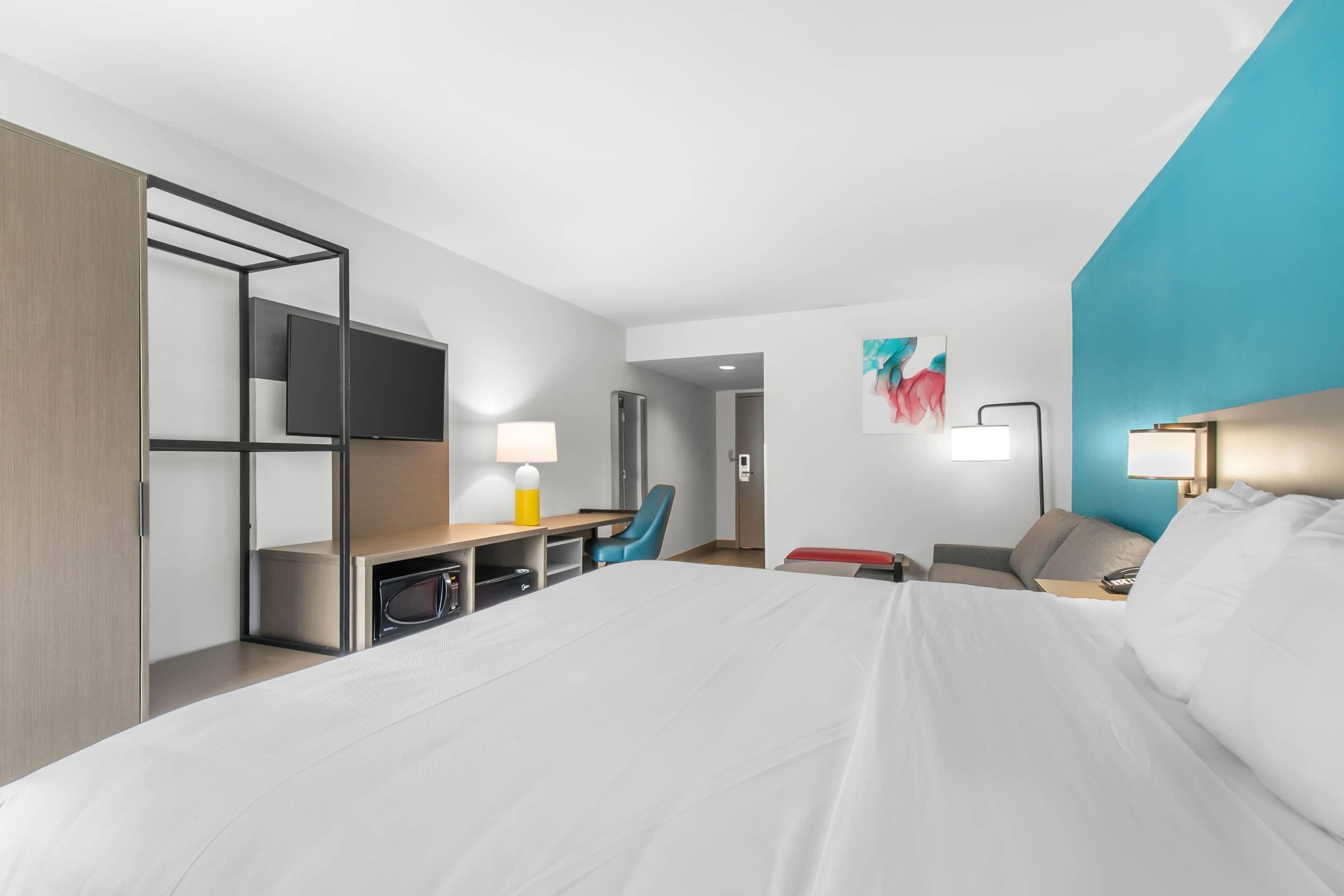 A modern hotel room with a large bed, a desk with a chair, a TV, and a lamp.