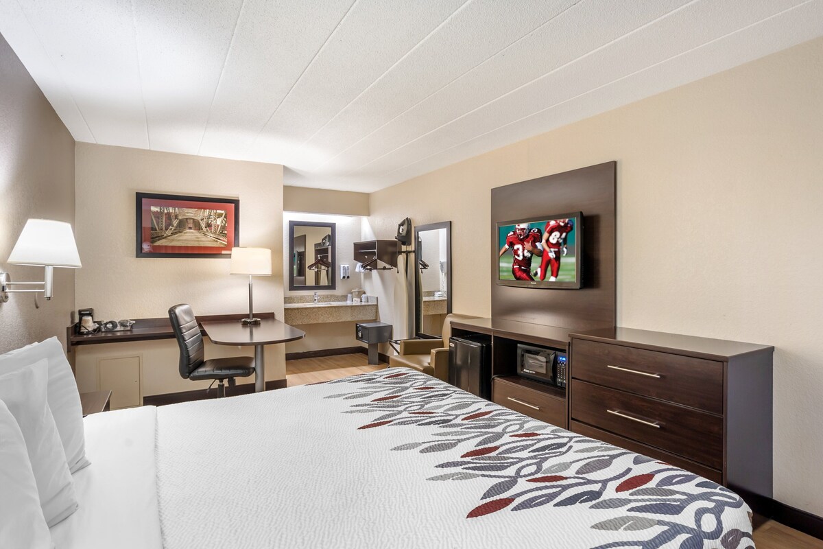 A hotel room with a large bed, a desk with a chair, a television, a mirror, and a minibar.