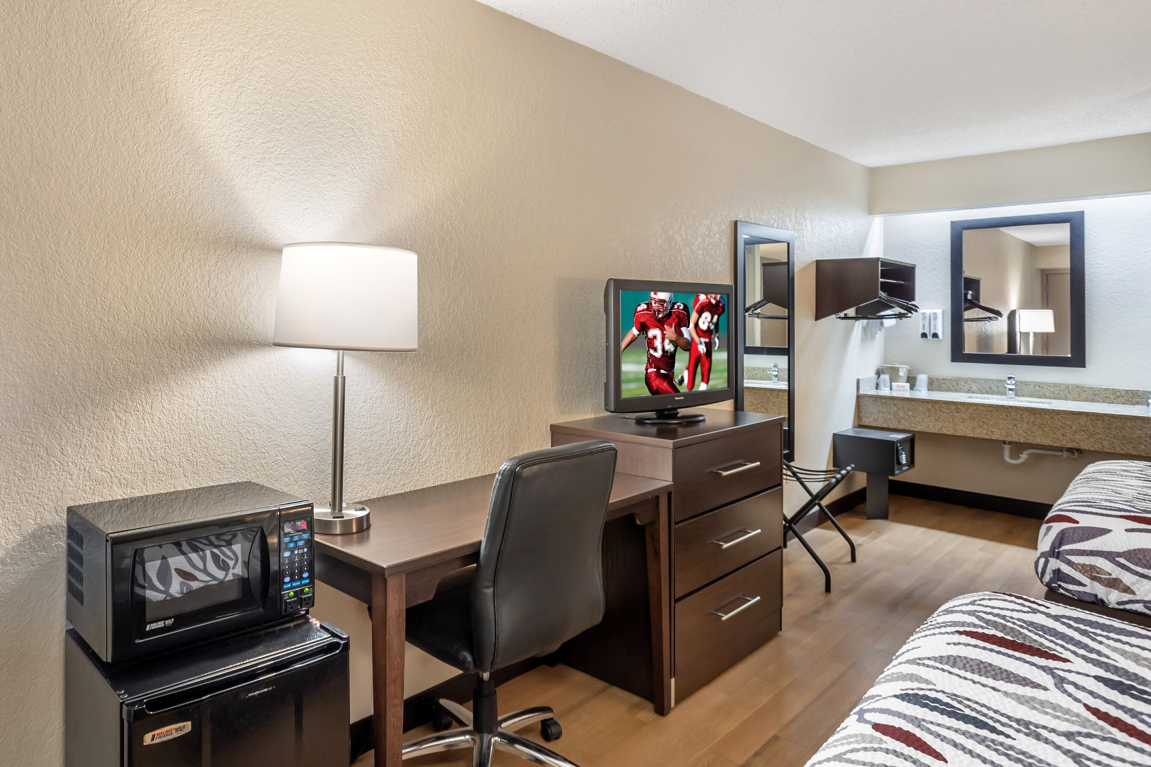 A hotel room with a desk, a television, a microwave, a chair, a bed, a dresser, a mirror, and a bathroom with a sink and towel rack.