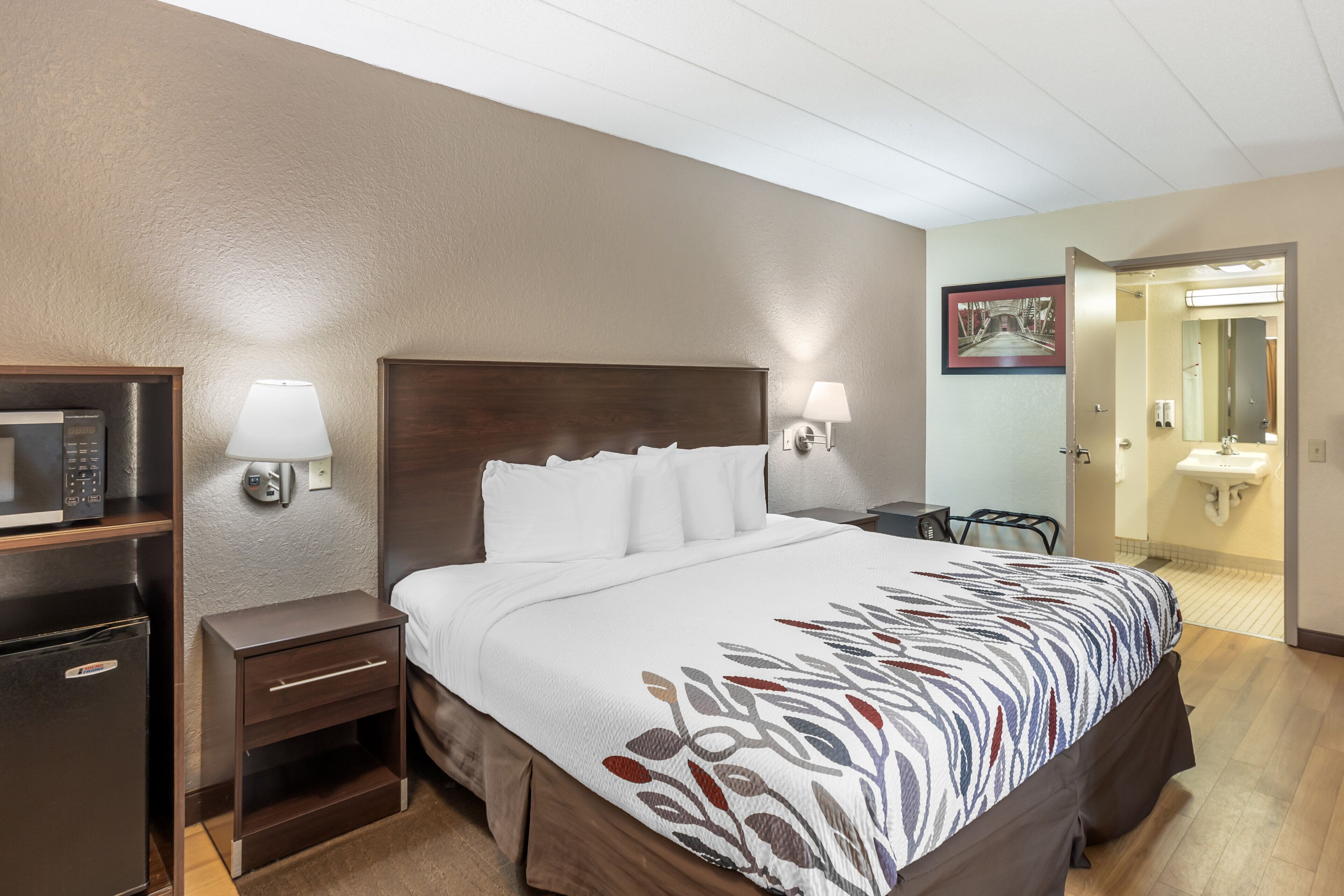 A hotel room with a bed, a nightstand, a lamp, a microwave, and a door leading to the bathroom.