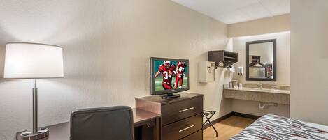 A hotel room with a bed, a desk with a chair, a television, a mirror, and a bathroom with a sink and mirror.