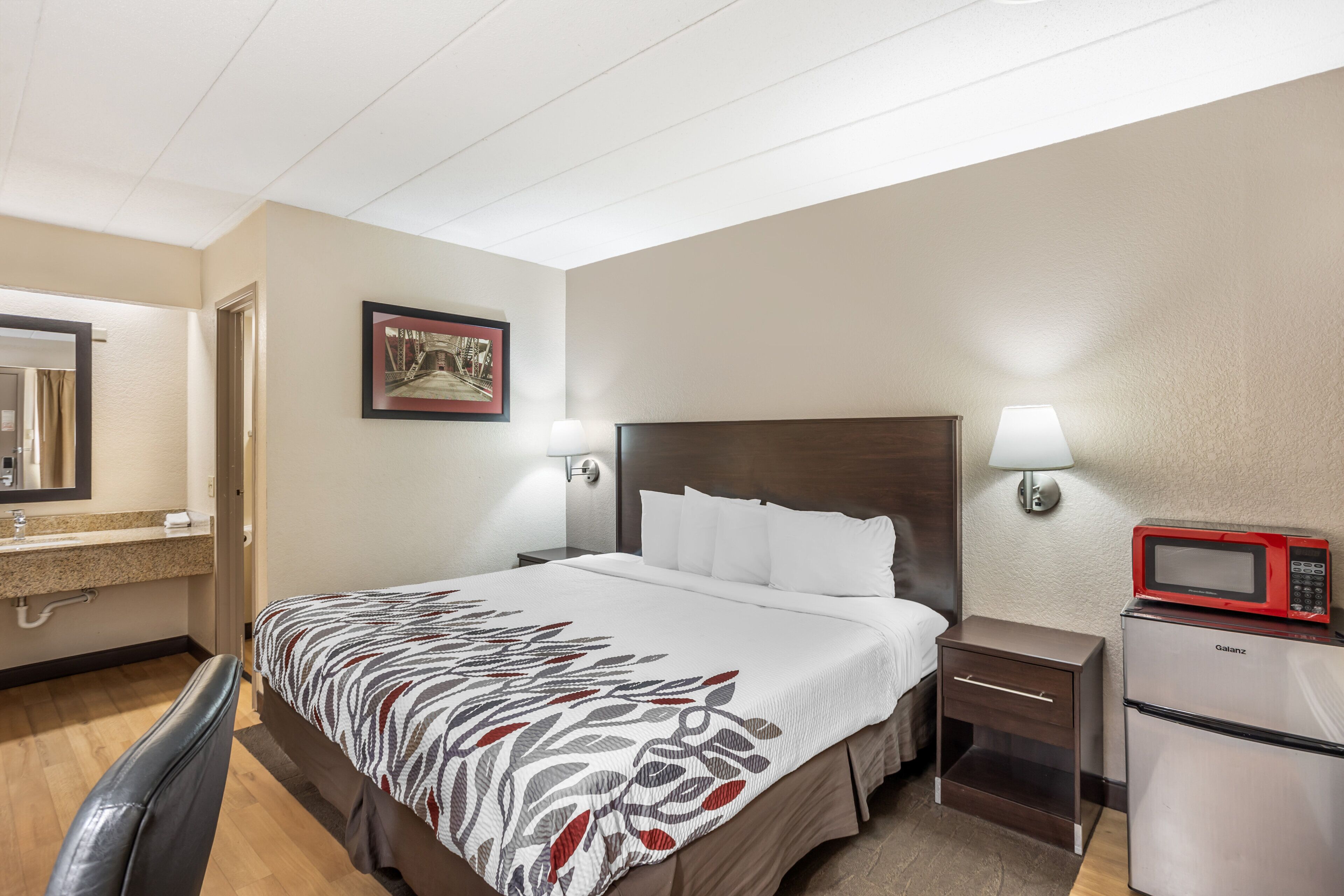 A hotel room with a bed, bedside tables, a microwave, a nightstand, a wall-mounted lamp, a framed picture, and a bathroom with a sink and mirror.