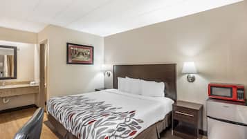 A hotel room with a bed, bedside tables, a microwave, a nightstand, a wall-mounted lamp, a framed picture, and a bathroom with a sink and mirror.