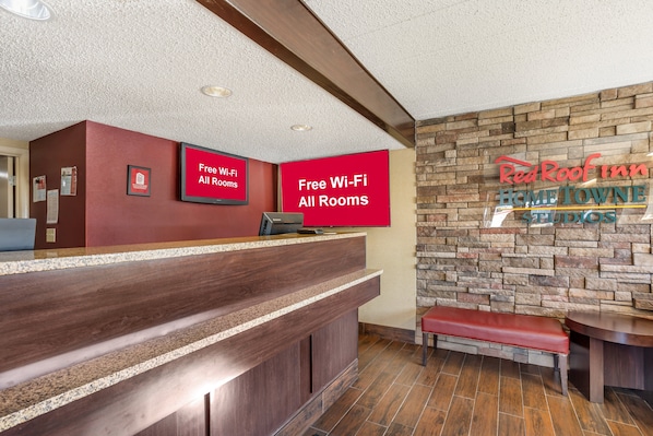 A hotel reception area with a wooden reception desk, a red wall with a 'Free Wi-Fi All Rooms' sign, and a stone wall with 'Red Roof Inn Home TOWNE Studios' branding. - Red Roof Inn Danville, PA (Danville)