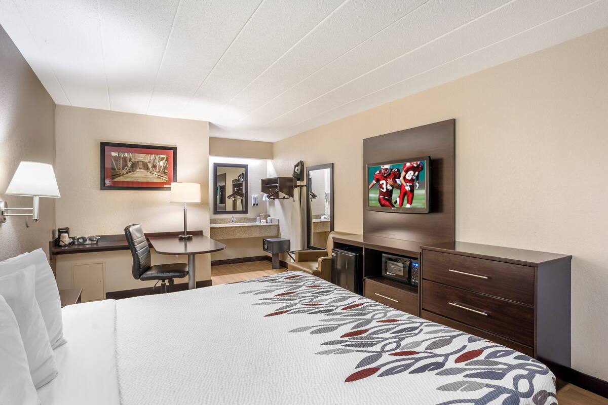 A hotel room with a large bed, a desk with a chair, a television, a mirror, and a minibar.