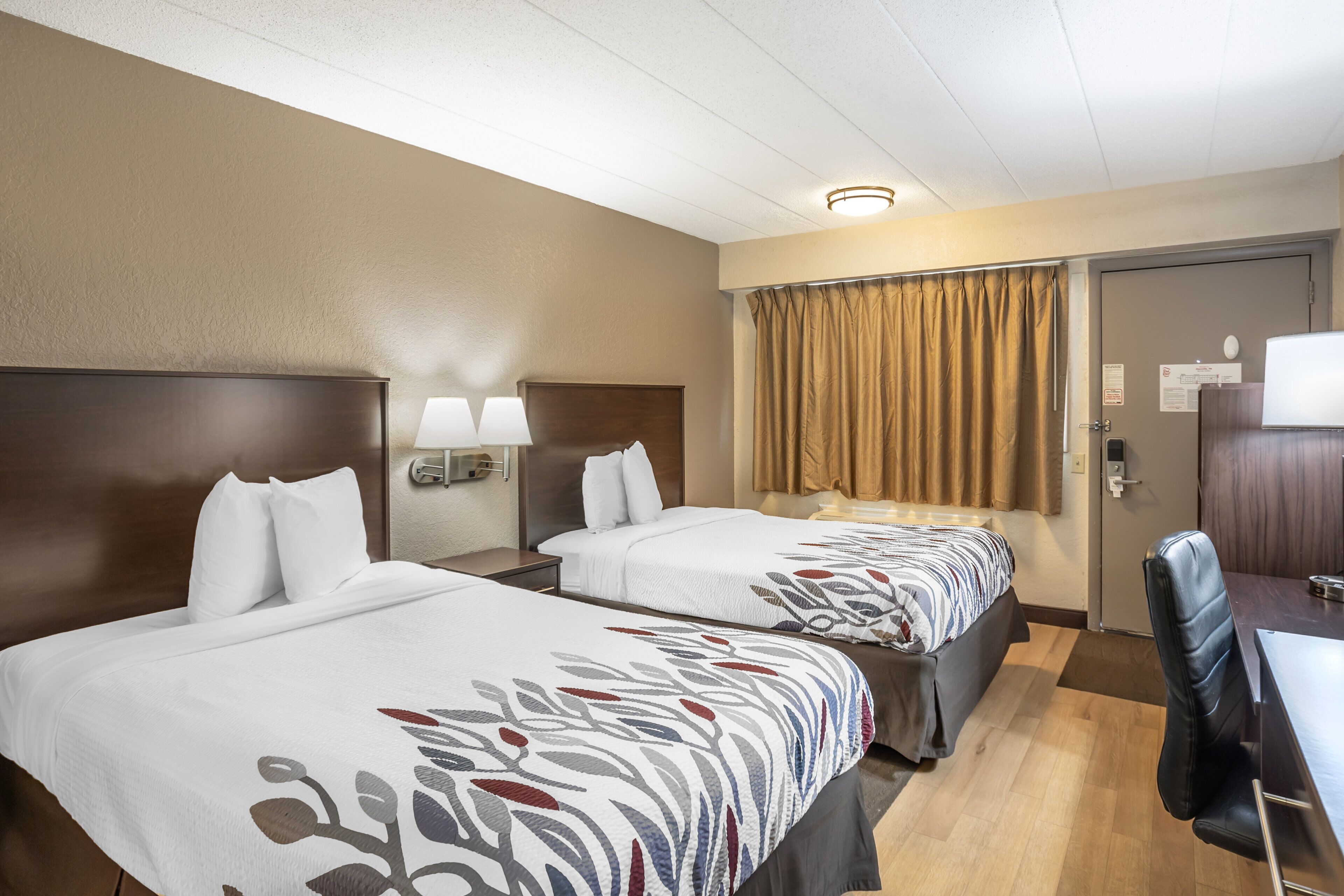 Ett hotellrum med två sängar, ett skrivbord, en stol, en lampa, en gardin och en dörr.