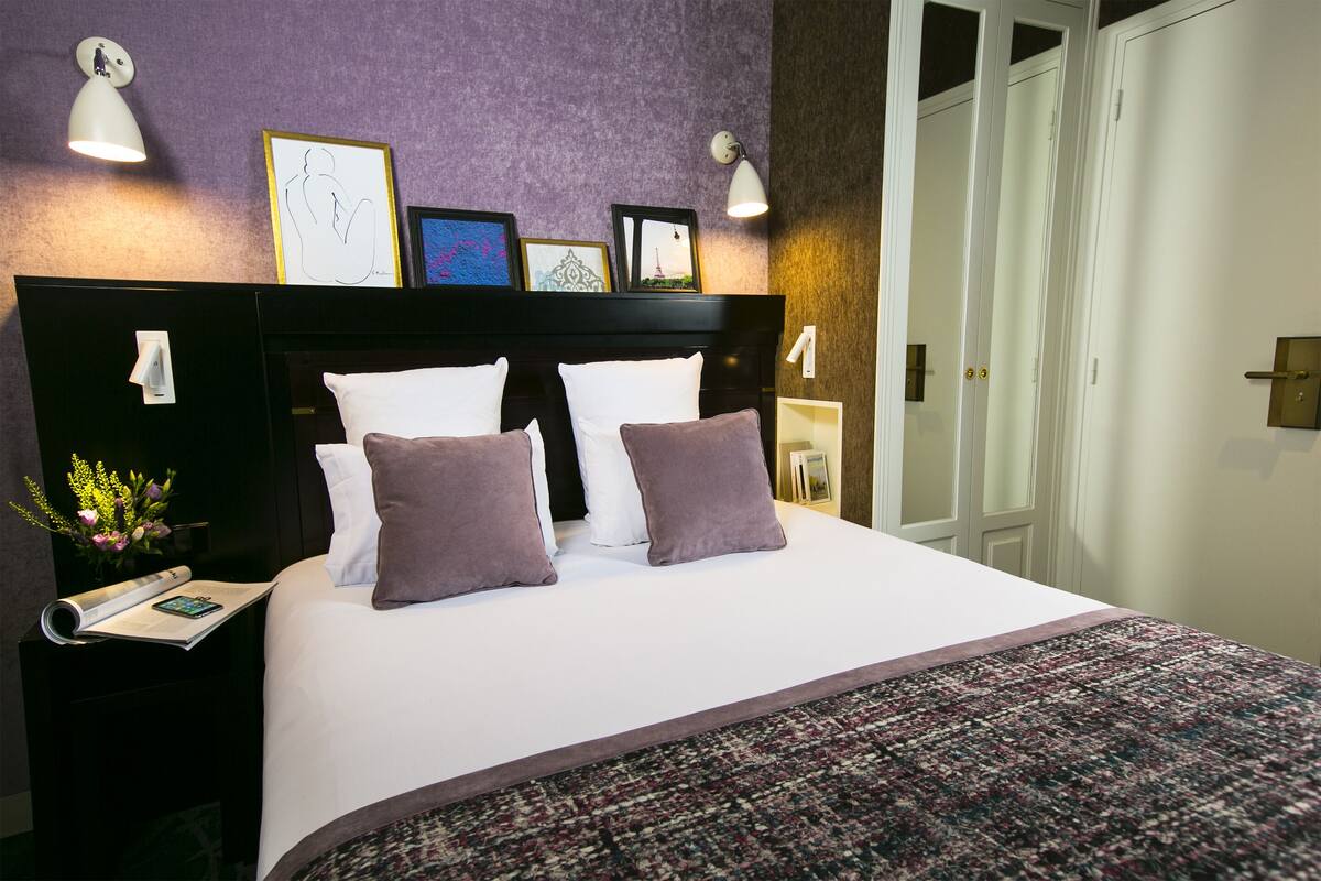A neatly made bed with white and purple bedding, a patterned rug, and framed pictures on the headboard.