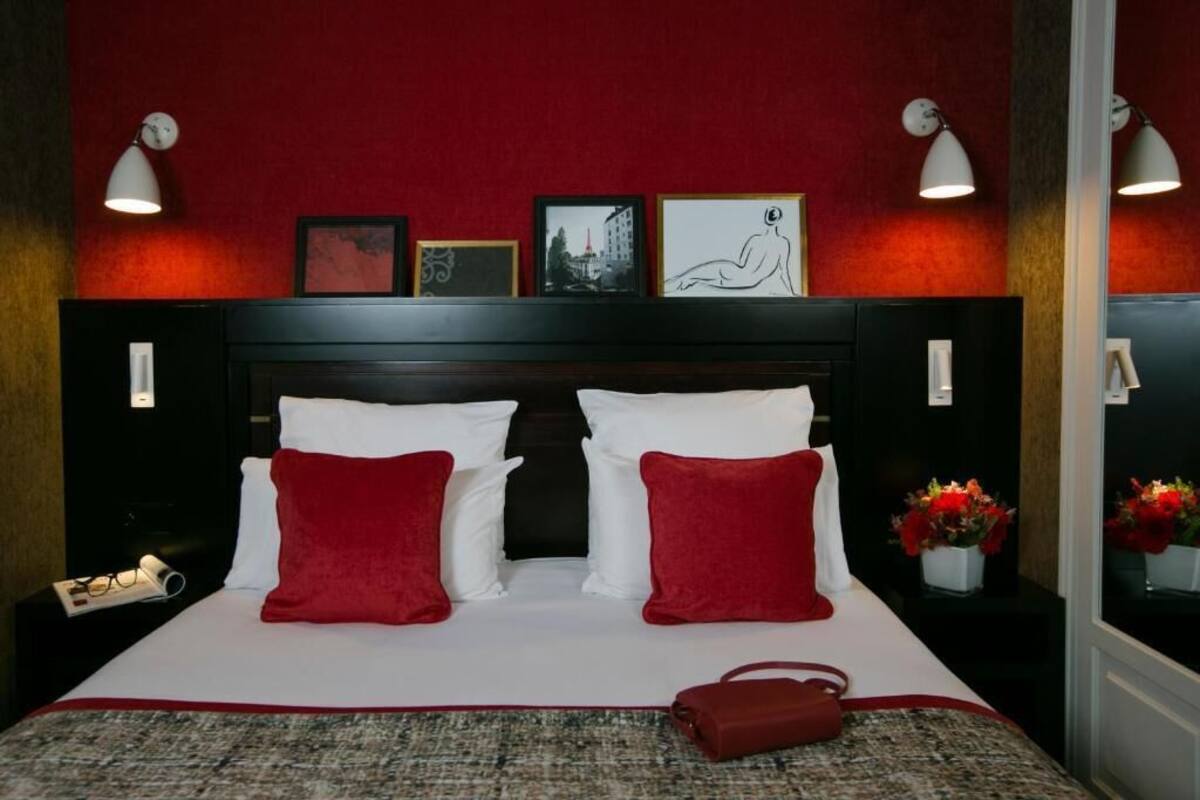 A hotel room with a bed, red and white pillows, a red handbag, framed pictures on the wall, and a nightstand with a lamp.