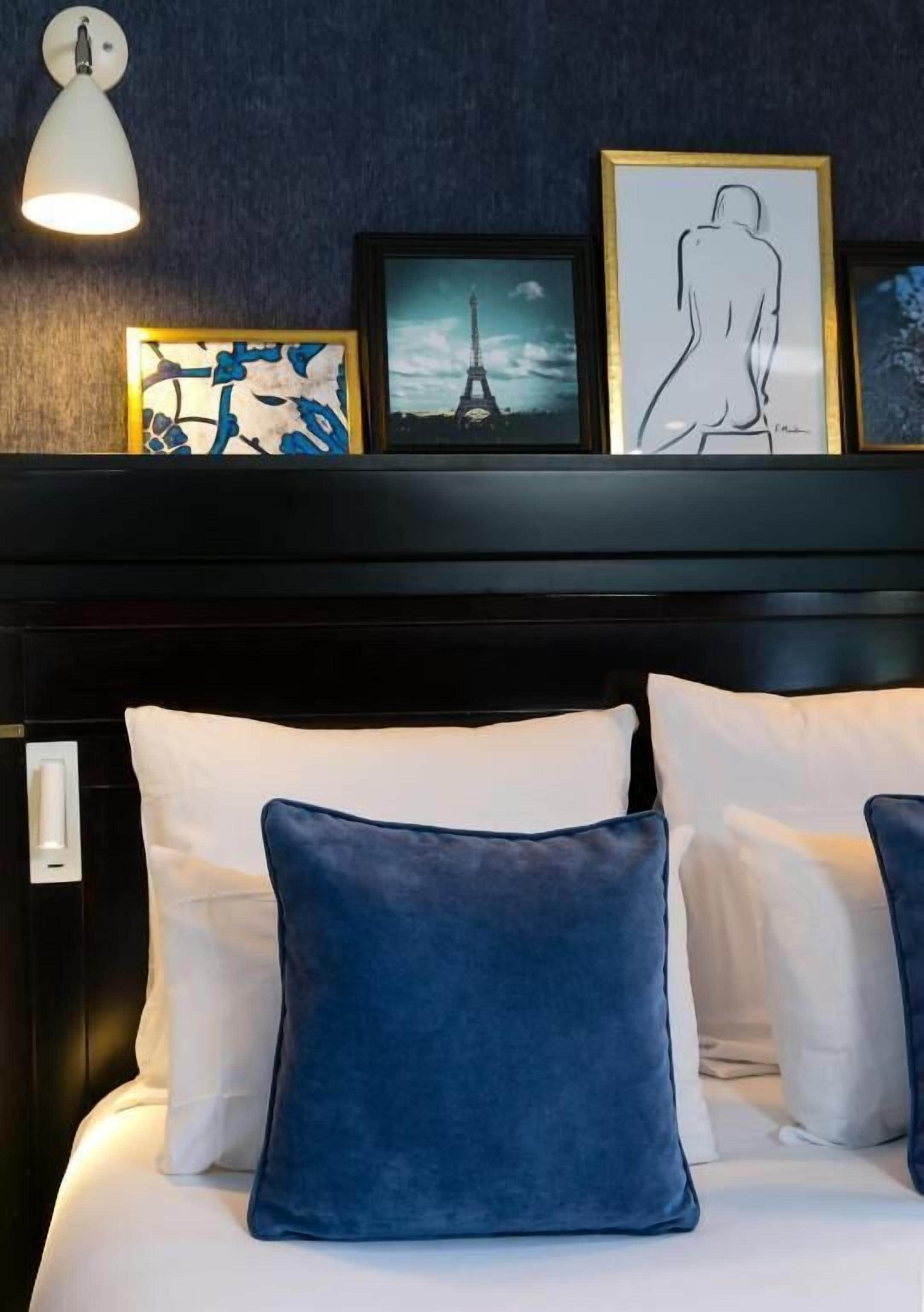 A bed with white and blue pillows, a wall-mounted light, and framed artwork including a silhouette and a cityscape.