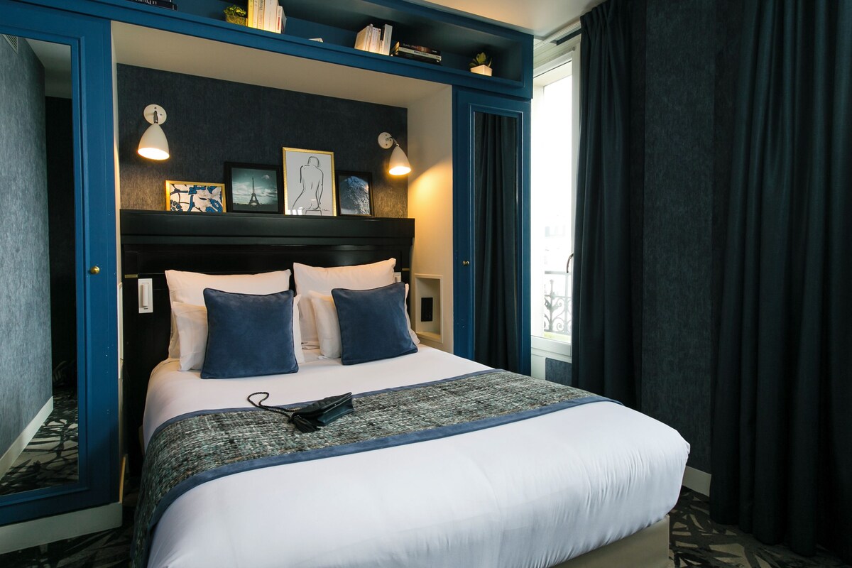 A hotel room with a bed, a nightstand, a wall-mounted shelf with books, and a window with curtains.