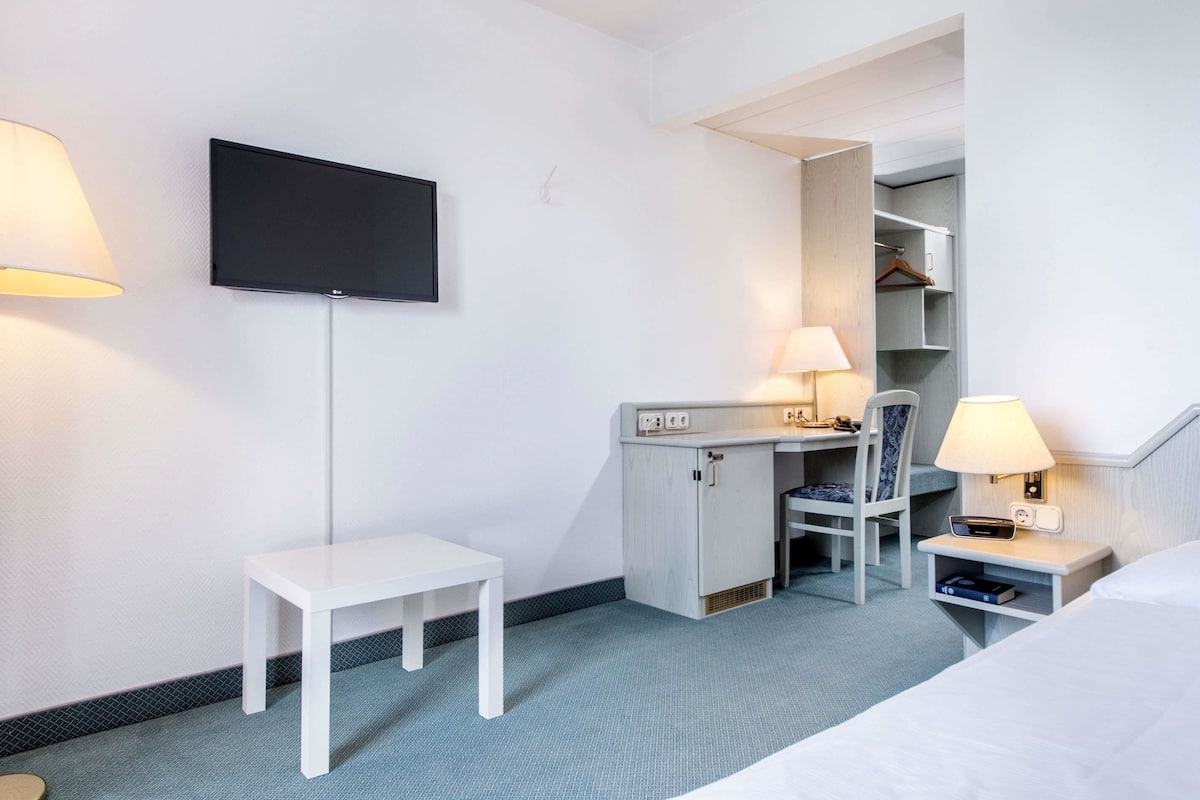 A hotel room with a bed, a desk with a chair, a TV mounted on the wall, and a wardrobe.