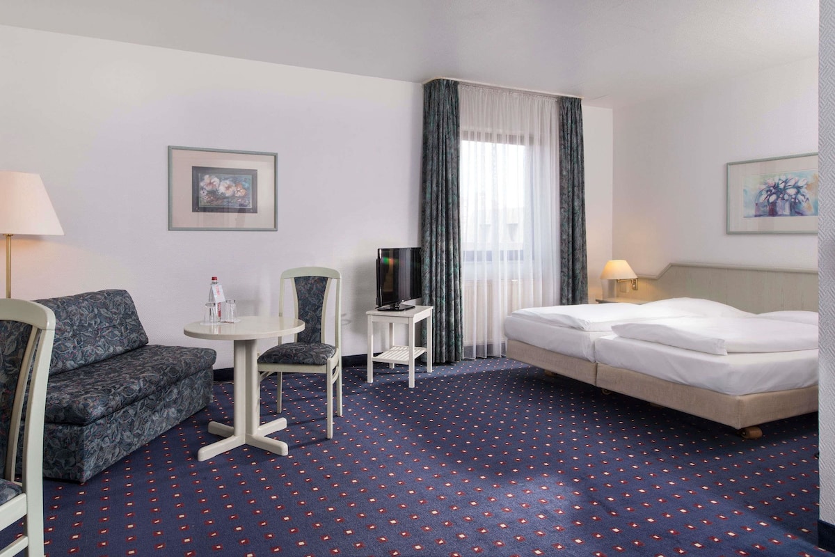 A hotel room with a bed, a sofa, a chair, a small table, a TV, and a lamp.