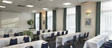 Un salón de conferencias con filas de mesas y sillas, ventanas grandes con cortinas y una pantalla de proyector en la pared.