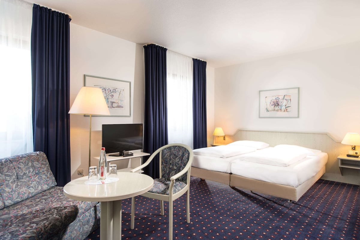 A hotel room with a bed, a sofa, a small table, a chair, a TV, and two wall-mounted lamps.