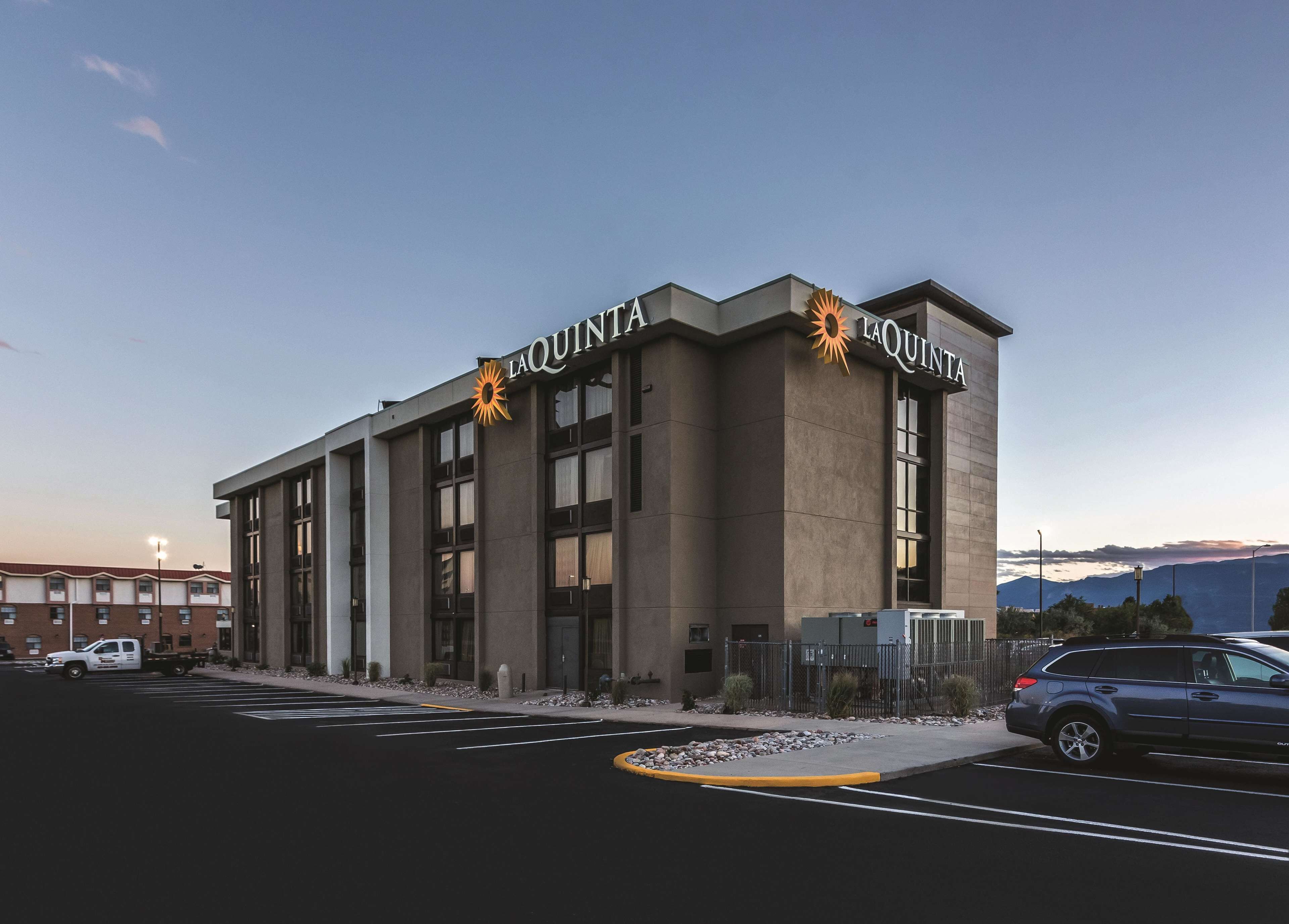 A modern building with the name "LAQUINTA" prominently displayed. It features a large entrance with a glass facade, multiple windows, and a parking lot in front.