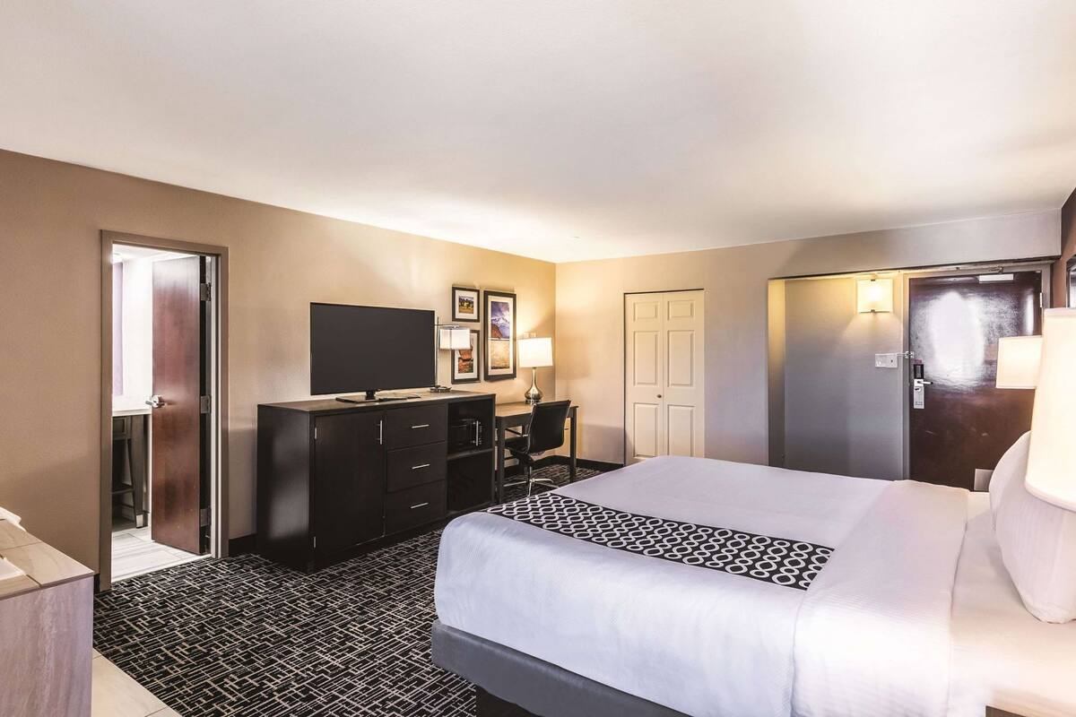 A hotel room with a bed, a desk with a TV, a dresser, and a door.