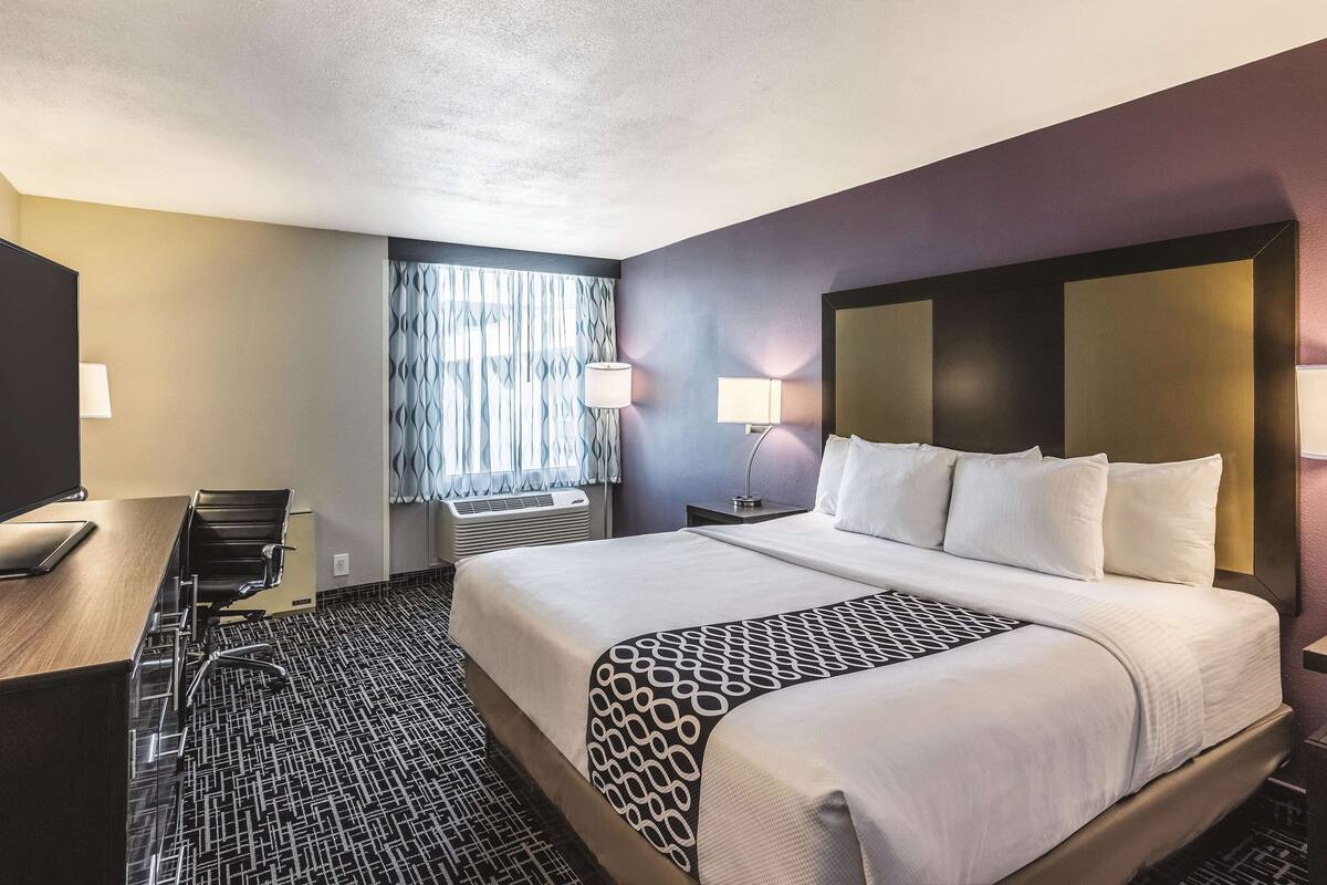 A hotel room with a bed, a desk, a chair, a lamp, and a window with curtains.